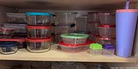 Cupboard shelf showing multiple stacked glass and plastic containers with colored lids and tall tumbler at right