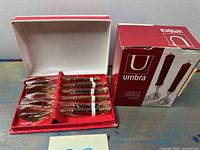 open spoon box showing six wrapped gold-tone spoons beside Umbra candle holder box