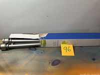 Curtain rod partially visible inside damaged blue and white box with label, showing silver metal rods