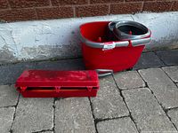 Red spin mop bucket beside closed red case on stone paving