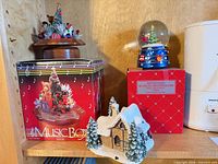 All three holiday items on shelf: tree music box, boxed snow globe, boxed music box, cottage figurine