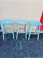 Front view of three turquoise wooden stools on pavement
