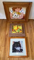 All three framed artworks standing on floor: woman with dog painting, floral painting, black cat print
