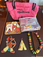 Lot overview showing pink Philips Sonicare tote, sealed bag of puzzle pieces, wooden cat puzzle, archer and teepee wooden pieces, wooden bead necklace