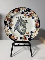 Front view of porcelain plate showing floral border, anatomical heart graphic, displayed on stand (stand not included)