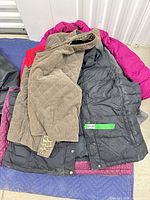 Pile of four outerwear pieces showing tan corduroy vest, black puffer vest, fuchsia jacket, hint of red vest