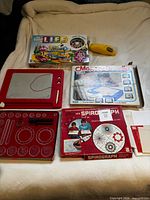 Overall lot showing Spirograph set, Magic-Doodle board, Game of Life box, yellow spinner, papers