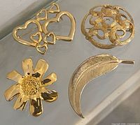 Four gold-tone brooches laid out: hearts, swirl, flower, feather