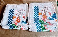 Stack of folded white cotton terry tea towels showing geese design and blue checkerboard border