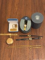 All items laid out on wooden surface including pocket watch, chain, two leather-strap watches, mesh watch band, Citizen watch in case with booklet, belt buckle and money clip in blue box