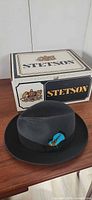 Black Borsalino fedora with blue feather resting on wooden surface in front of Stetson box