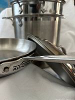 Close-up All-Clad logo on fry pan handle with stock pot and steamer in background