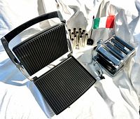 All three main items displayed together: panini grill open, pasta machine with crank and clamp, 12 cannoli tubes beside Italian flag (flag not included)