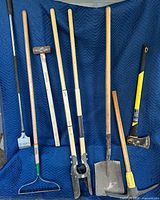 Eight long-handle yard tools including scraper, sledgehammer, bow rake, post hole digger, square shovel, pick mattock, wood splitting axe displayed upright
