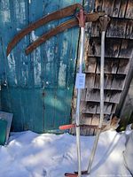 Both aluminum scythes showing full length shafts, handgrips and rusted blades