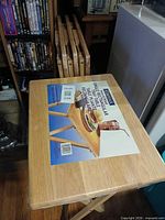 Top view of one Cosco wood folding TV tray table with label and plastic wrap, stack of additional tables in background