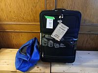 Large black upright suitcase with packaging and attached tags, blue tote beside