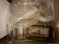 Box containing assorted clear glass bakeware pieces including round casserole lid, round handled bowl, pie plate, two square dishes, rectangular loaf dish, some wrapped in bubble wrap