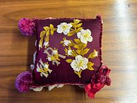 Front view of burgundy pillow with floral embroidery and pom-poms