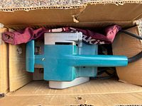 Top view of teal Makita 9900B belt sander and maroon dust bag inside box