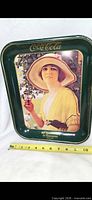 Front of green Coca-Cola tray showing graphic of woman holding drink and measuring tape for scale