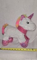 Side view of unicorn plush showing body, horn, mane, tail, scarf, hooves, and measuring tape for scale