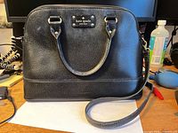 Front view of black pebbled leather Kate Spade satchel showing handles, logo plate, detachable shoulder strap