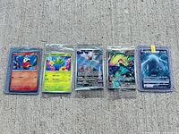 Five individually sealed Pokémon trading cards displayed in protective sleeves and top-loaders on concrete surface