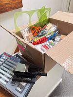 Box with various sealed office supplies including templates, scissors, pens, mounting squares
