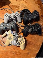 Six Sony wired PlayStation controllers in grey, black and beige with attached cables