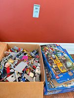 Open cardboard carton filled with assorted loose LEGO pieces; 60160 Jungle Mobile Lab box leaning at side
