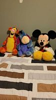 Group shot of Tigger, Eeyore and Mickey plush sitting on bed