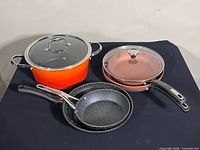 Overall view of cookware set including orange pot, copper sauté pan with lid, two speckled skillets