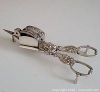 Side view of ornate silver-plated candle snuffer showing handles, feet, and wick box