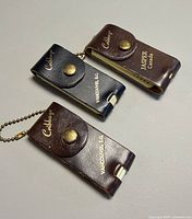 Three leather-covered miniature cribbage board keychains closed, showing gold lettering and ball chains