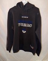 Full view of black BOSS x 76ers hoodie on hanger, showing front graphics and sleeve stripes