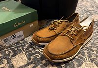 Pair of tan Clarks Allston Edge shoes beside original green box on patterned carpet