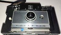 Front view of Polaroid Land Camera Automatic 100 seated in hard carrying case showing lens, rangefinder, strap, and interior of case.