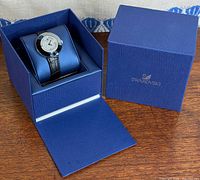 Watch seated on blue cushion within open Swarovski box next to matching lid