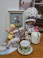 Group image showing entire lot of assorted vintage porcelain/ceramic children's figurines, animal figurines, teacup with saucer, cross-stitch artwork, cookie jar, and canister