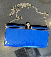 Front view of blue patent clutch with gold-tone chain displayed on dark surface