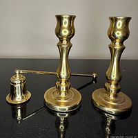 Two polished brass candlesticks and candle snuffer on black surface