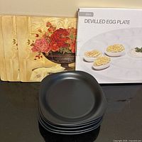 Stack of five black dishes, deviled egg plate box, three floral placemats visible