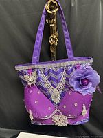 Front view of purple embellished shoulder bag hanging, showing handles and decorations