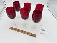 Six ruby red wine glasses with ruler and note card visible for scale and identification.