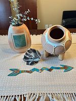 Group shot of ceramic vase with stems, terracotta pot on legs, painted stone slab, arrow-fish wall accent and small stones