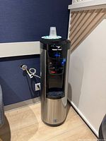 Full view of cylindrical stainless-steel water cooler showing control panel, taps, and attached plumbing line