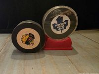 Front view of two hockey pucks: autographed Toronto Maple Leafs puck in case on red stand; Chicago Blackhawks logo puck