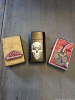 Front view of three Zippo lighters: Budweiser 125th, skull graphic, Libra zodiac