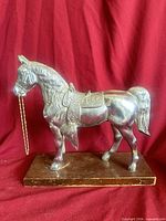 Front left view of silver-tone metal horse on wood base showing saddle and chain bridle